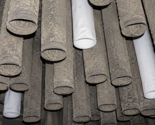 Inside an industrial baghouse showing mostly dirty filter bags with three new replacement filter bags installed among them during maintenance.